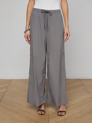 A person in loose-fitting, gray Valkyrie Snap Pant with a white top, black open-toe heels, and a bracelet stands on a wooden floor against a light gray wall.