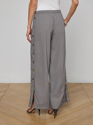 A person is photographed from the back, standing on a wooden floor, wearing loose gray Valkyrie Snap Pant with large side buttons, styled with a white top and heeled sandals.