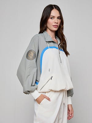 A woman with long brown hair wears the Aurelia Windbreaker—a loose, light gray and white retro jacket with blue accents and a round sleeve patch. She stands facing the camera, one hand in her pocket, against a plain light background.