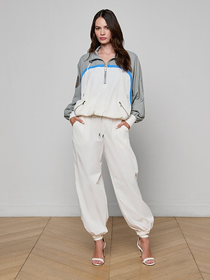 A woman stands against a plain wall in the Aurelia Windbreaker, a loose, light-colored athleisure tracksuit with gray and blue accents, hands in pockets and wearing white high-heeled sandals on a wooden floor.