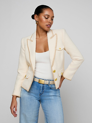 A woman poses confidently in the Wylla Tweed Blazer—an ecru jacket with gold buttons and embroidered trim—styled with a white tank top, light blue jeans, beige belt, gold hoop earrings, and a sleek bun against a plain background.