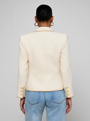 A person with hair in a neat bun is shown from behind, wearing the Wylla Tweed Blazer with embroidered trim and gold sleeve buttons, paired with light blue jeans and large hoop earrings.