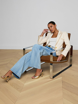 A woman sits in a modern brown armchair, wearing the Wylla Tweed Blazer over a white top, with light blue flared jeans and gold heels. She rests her elbow on the armrest, gazing thoughtfully at the camera in a minimal, uncluttered setting.