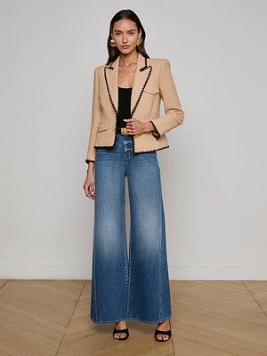 A woman stands in a neutral room wearing the Wylla Tweed Blazer by L'AGENCE Sale, styled with a black top, wide-leg blue jeans, and black heeled sandals. Her chic look and earrings highlight the elegance of classic neutrals.