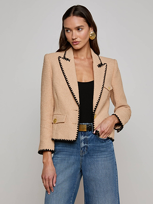 A woman in the Wylla Tweed Blazer by L'AGENCE Sale, styled with a black top and blue jeans, stands against a plain light background. She has long brown hair, looks to the side, and keeps one hand in her pocket.