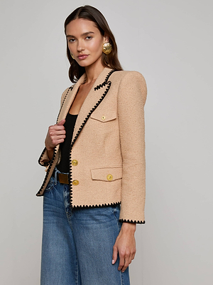 A woman wearing the L'AGENCE Sale Wylla Tweed Blazer, featuring neutral tan tweed with embroidered trim and gold buttons, pairs it with a black top and blue jeans as she stands against a plain gray background, smiling slightly at the camera.