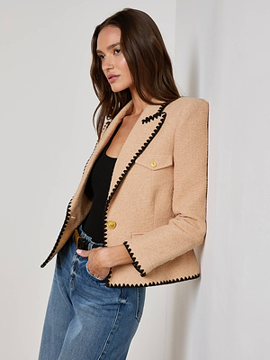 A woman with long brown hair, wearing the Wylla Tweed Blazer by L'AGENCE Sale over a black top and blue jeans, leans against a white wall and looks to the side with a neutral expression.