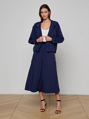 A woman stands indoors on a wooden floor in a navy blue Geovana Hooded Windbreaker and matching midi skirt over a white top, paired with black high-heeled sandals. Facing the camera with a neutral look, she embodies effortless travel style.