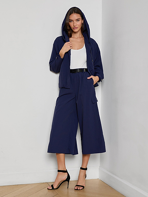 A woman stands indoors wearing a navy blue Geovana Hooded Windbreaker, matching wide-leg culottes, a white top, a black belt, and black high-heeled sandals. One hand is in her pocket as she looks at the camera.