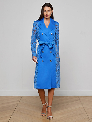 A woman stands on a wooden floor against a plain light wall, wearing the Amata Lace Trench Coat in regatta blue with floral lace sleeves and side panels, paired with silver strappy high-heeled sandals.