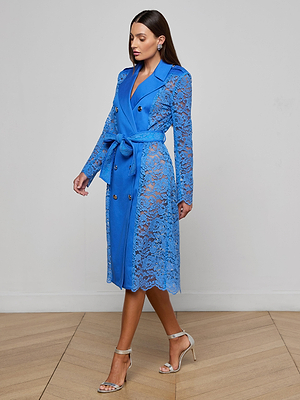A woman poses indoors on a wooden floor wearing the Amata Lace Trench Coat in regatta blue, featuring sheer floral lace, a waist tie, and gold buttons. She pairs it with silver high-heeled sandals and styled hair.