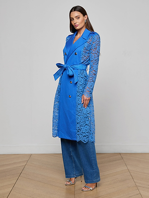 A woman stands indoors in the Amata Lace Trench Coat in regatta blue, featuring a double-breasted floral lace design and matching belt, styled with blue wide-leg jeans and silver high-heeled sandals against a plain, neutral background.
