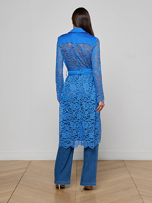 A woman with straight, dark hair faces away, wearing the Amata Lace Trench Coat in regatta blue with a floral lace pattern, belted over blue jeans and heeled shoes. She stands on a wooden floor against a plain wall.