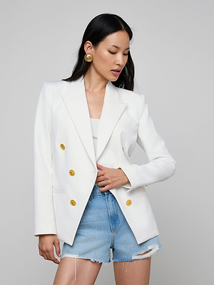 A woman with long black hair wears the Kyomi Double-Breasted Blazer in white over a light top and distressed denim shorts, looking to the side with one hand on her blazer against a plain background.