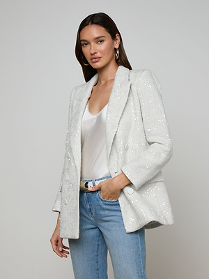 A woman stands against a plain light background with one hand in her pocket, wearing the Cambrie Oversized Sequin Blazer over a white top and light blue jeans, her expression neutral.