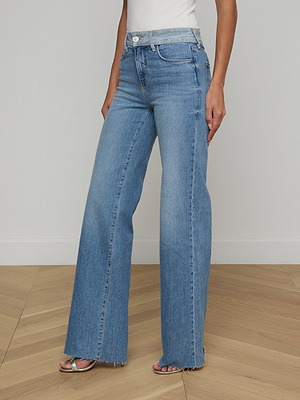 A woman wears the Louane Wide-Leg Flare Jean in light blue stretch denim with a raw hem, styled with a white sleeveless top and nude heeled sandals. She stands on a light wooden floor against a plain white wall.