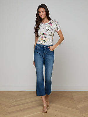 A woman with long brown hair and a relaxed expression stands on a wooden floor, wearing a white floral short-sleeve top, beige heeled sandals, and the Kendra Cropped Flare Jean in dark denim. The background is a plain light wall.