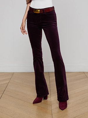 A person wears the L'AGENCE Stevie Velvet Jean in deep purple, a white top, a red belt with a gold buckle, and matching purple pointed-toe shoes while standing on a wooden floor against a light gray wall.