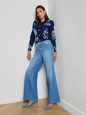 A woman with long brown hair and a confident expression stands against a white wall, wearing the Alicent Wide-Leg Jean in lightweight denim, paired with a blue floral jacket and blue pointed-toe heels.