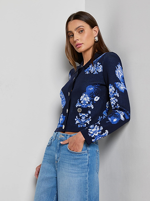 A woman with straight brown hair models the Alicent Wide-Leg Jean and a navy floral jacket with silver buttons, standing against a white wall and looking confidently at the camera.