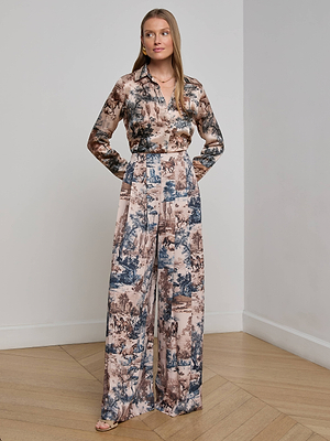 A woman poses confidently indoors, wearing the Nova Pleated Wide-Leg Pant—a wide-leg trouser set with a scenic Western toile print in beige, blue, and brown—paired with a matching blouse.