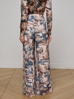 A person stands on a wooden floor, seen from the back, wearing Nova Pleated Wide-Leg Pant and a matching shirt in blue and brown fluid suiting fabric with a Western toile print of pastoral scenes.