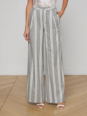 A person wearing the Allanah Striped Trouser—modern, wide-leg gray pants in an Italian wool blend with white vertical stripes—paired with a white sleeveless top and heeled sandals stands on a wooden floor against a plain wall.