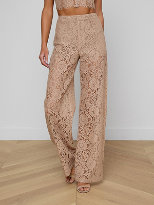 A woman stands on a light wooden floor against a plain white wall, wearing the Livvy Lace Trouser—a high-rise, wide-leg cappuccino lace pant—paired with a matching lace crop top and open-toe heels.