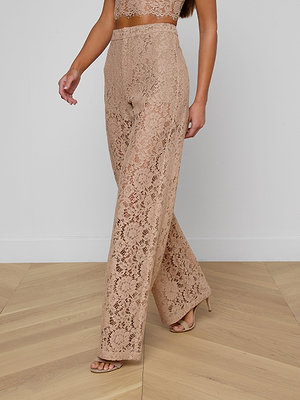 A woman stands on a light wooden floor against a white wall, wearing the Livvy Lace Trouser in a high-rise, wide-leg cappuccino style with a matching crop top and nude heeled sandals.