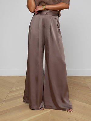 A woman in the Aubrie Satin Pant—high-waisted, wide leg with pleats—paired with a cardamon brown top and gold cuff bracelet stands on a light wood floor against a white wall.