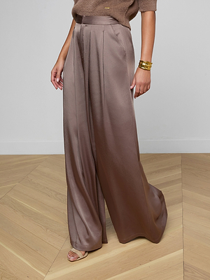 Wearing the Aubrie Satin Pant in cardamon brown with front pleats, a taupe short-sleeve sweater, gold bracelets, and beige heeled sandals, a person stands on light wood flooring.
