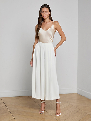 A woman with long, wavy brown hair stands indoors wearing a sleeveless light beige satin top, the Novalynn Cropped Pleated Pant in white, and white high-heeled sandals.