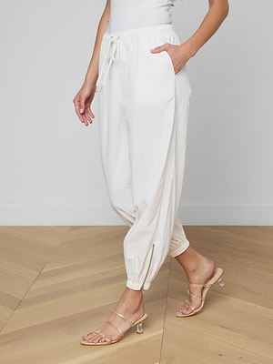 A person wears the Mariko Jogger in modern ivory, crafted from water-repellent nylon, paired with beige high-heeled sandals, standing on a wooden floor against a plain light-colored wall.