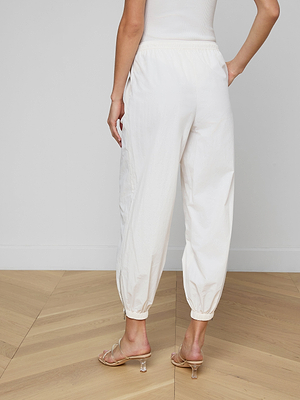 Wearing the Mariko Jogger in ivory and nude high heels, a person stands on a wooden floor facing away from the camera. The modern design of the joggers stands out against a plain white wall, creating a clean, minimalist look.