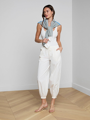 A woman stands indoors on a wooden floor wearing the Mariko Jogger, an ivory sleeveless top, nude sandals, and a light gray sweater draped over her shoulders. One hand is in her pocket as she looks to the side.
