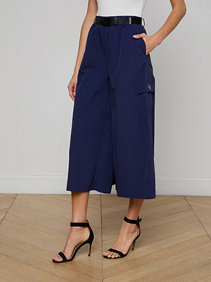 Wearing the Shoda Cropped Wide-Leg Pant in navy with a black belt, white sleeveless top, and black high-heeled sandals, this person stands on a wooden floor with hands in pockets.