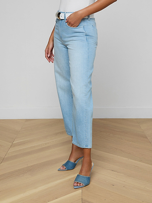 Wearing the Mallis Bow-Leg Jean in light wash with a white belt and blue heeled sandals, a person stands on a light wood floor against a plain wall. Only the lower half of their body is shown.