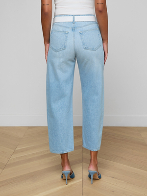 A person is shown from the back standing on a wooden floor with a white wall, wearing Mallis Bow-Leg Jean in a light wash and heeled sandals.