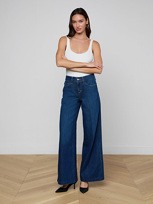 A woman with long dark hair stands indoors against a plain wall, wearing a white tank top, black heels, and the Pacey Wide-Leg Jean. Her arms are crossed.