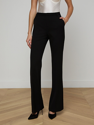 Wearing the Frances Bootcut Trouser in black, a white satin top, and black pointed-toe heels, a person stands on a wooden floor with one hand in their pocket. The upper part of their face is not visible.