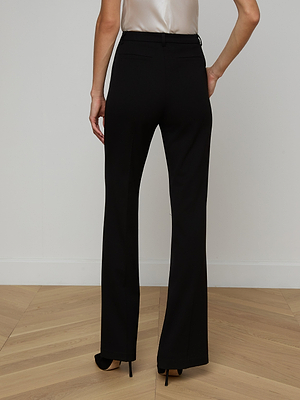 Wearing the Frances Bootcut Trouser in stretch suit fabric and black heels, a person stands on a light wood floor before a plain white wall—a timeless wardrobe staple.