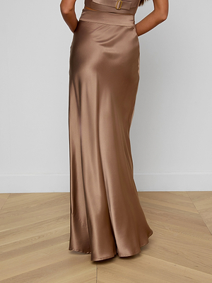 A woman stands on a light wooden floor, photographed from behind against a plain white wall, wearing the Ryker Silk Maxi Skirt—a shiny, floor-length, bias-cut silk skirt with a fitted waist.