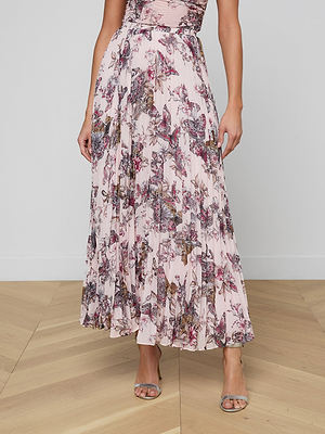 A person wearing the Torin Pleated Maxi Skirt—an ankle-length poly chiffon skirt in light pink with pleats and pink-purple hues—paired with silver high-heeled sandals, stands on a wooden floor against a plain white wall.