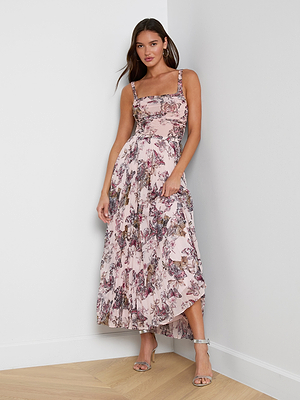 A woman stands against a white wall wearing the Torin Pleated Maxi Skirt, a long, light pink poly chiffon skirt with a floral print, paired with silver high-heeled sandals.