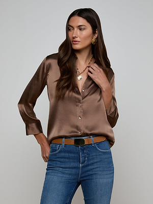 A woman with long brown hair wears the Dani Silk Blouse in cardamom brown, paired with blue jeans, a tan belt, and gold jewelry, standing against a plain light gray background.