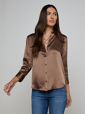 A woman with long brown hair wears the Dani Silk Blouse in shiny cardamom brown, styled as a button-down and paired with blue jeans. She stands against a plain light background, looking slightly to the side with one arm relaxed.
