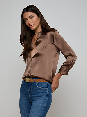 A woman with long brown hair wears a cardamom brown Dani Silk Blouse and blue jeans, posing with one hand on her hip against a plain gray background.