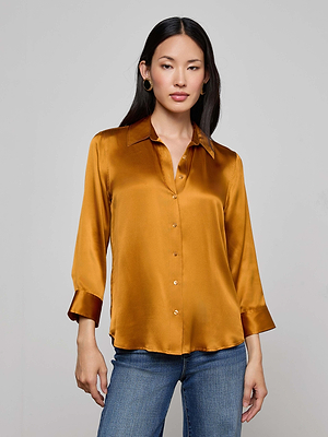 A woman with long dark hair models the L'AGENCE Dani Silk Blouse—a shiny gold, collared top with three-quarter sleeves—paired with blue jeans against a plain light gray background.