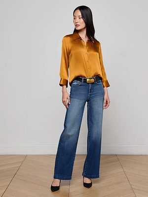 A woman poses indoors against a light background, wearing the L'AGENCE Dani Silk Blouse in gold with three-quarter sleeves, paired with blue high-waisted wide-leg jeans and black heels, one hand in her pocket as she looks to the side.