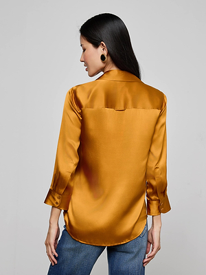 A woman with long dark hair is seen from behind wearing the L'AGENCE Dani Silk Blouse in shiny gold with long sleeves, paired with blue jeans, set against a plain light gray background.